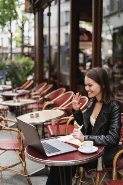 Siyah ceketli mutlu serbest çalışan Fransız açık hava kafesinde kahve masasının yanında video görüşmesi yapıyor. 