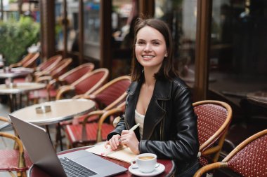 Siyah ceketli mutlu bir kadın laptopun yanındaki kameraya ve açık hava kafedeki masada bir fincan kahveye bakıyor.