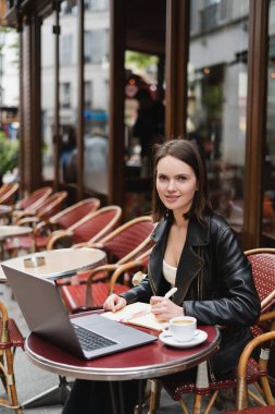Siyah ceketli neşeli bir kadın laptopun yanındaki kameraya ve açık hava kafedeki masada bir fincan kahveye bakıyor.