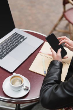 Siyah ceketli serbest çalışanın dizüstü bilgisayarın yanında akıllı telefon ve açık hava Fransız kahvesinde kahve tutuşu. 