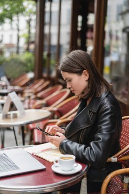 Siyah ceketli serbest çalışan bilgisayarın yanında akıllı telefon ve açık hava kahvesinde bir fincan kahve kullanıyor. 