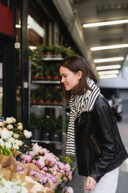side view of smiling young woman in stylish outfit choosing flowers on street in paris  clipart