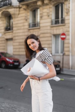 smiling young woman in stylish outfit holding bouquet wrapped in paper on street in paris  clipart