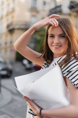 young woman in stylish outfit adjusting hair and holding bouquet wrapped in paper on street in paris  clipart