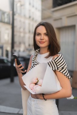 cheerful woman in trendy outfit holding smartphone and bouquet with peonies on street in paris clipart