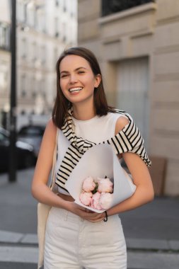 joyful and stylish woman holding bouquet of flowers wrapped in paper while standing on street in paris  clipart