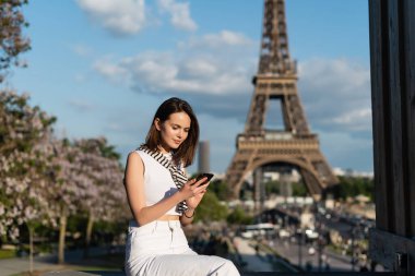 Paris 'teki Eyfel Kulesi' nin yanında otururken akıllı telefon kullanan şık kıyafetli genç bir kadın. 