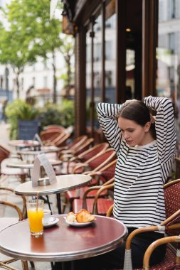 Çizgili uzun kollu gömlekli genç bir kadın içkilerin yanında saç düzeltiyor ve Paris 'teki açık kafede kruvasan yiyor.