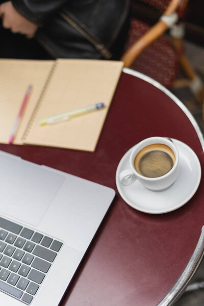 laptop and cup of coffee near blurred notebook on table in outdoor cafe 