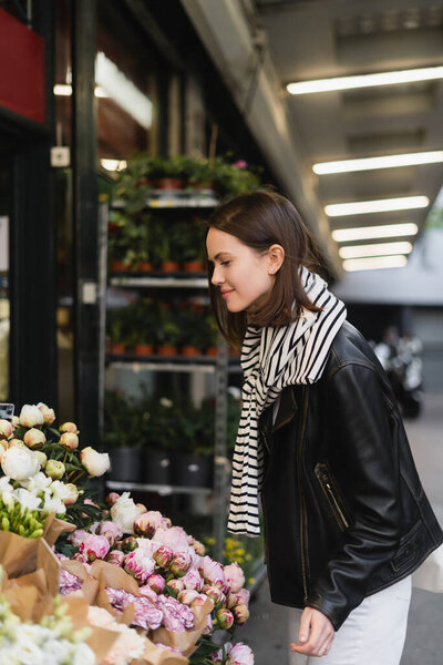 side view of smiling young woman in stylish outfit choosing flowers on street in paris 