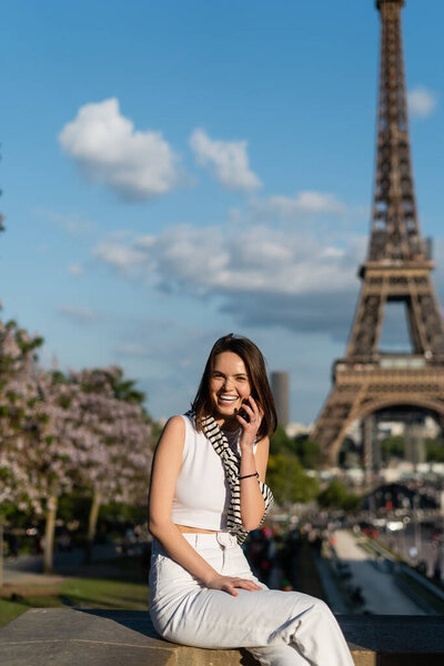 happy young woman in stylish outfit talking on smartphone while sitting near eiffel tower in paris, france