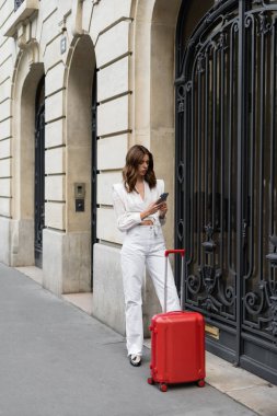 Fashionable traveler using cellphone near suitcase on street in Paris  clipart