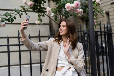 Positive and stylish woman having video call on smartphone near fence on street in Paris  clipart