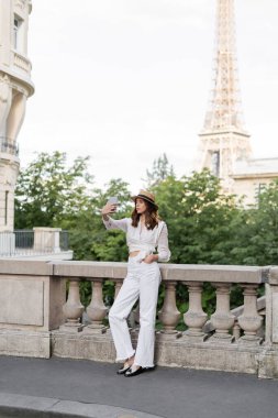 Paris 'teki Eyfel Kulesi' nin arka planında akıllı telefondan selfie çeken şık bir kadın.  