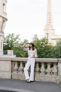 Fashionable woman taking selfie on smartphone with Eiffel tower at background in Paris   clipart
