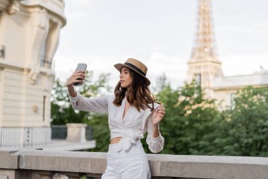 Güneş şapkalı turist Paris 'te Eyfel Kulesi' nin arka planında cep telefonuyla selfie çekiyor. 