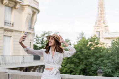 Güneş şapkalı pozitif gezgin Fransa 'da Eyfel Kulesi' nin arka planında akıllı telefonuyla selfie çekiyor. 