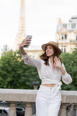 Güneşli şapkalı gülümseyen turist Paris 'teki Eyfel Kulesi' nin arka planında akıllı telefonuyla video görüşmesi yapıyor.