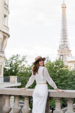Bluz ve güneş şapkalı mutlu genç kadın Paris 'te Eyfel Kulesi' nin arka planında duruyor. 