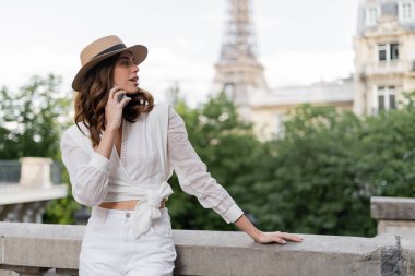 Güneşli şapkalı gülümseyen turist Paris 'te arka planda Eyfel Kulesi ile akıllı telefondan konuşuyor. 
