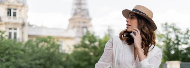 Woman in sun hat talking on smartphone with blurred Eiffel tower at background in Paris, banner  clipart