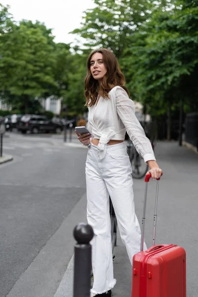 Smiling woman with suitcase and mobile phone standing near road in Paris