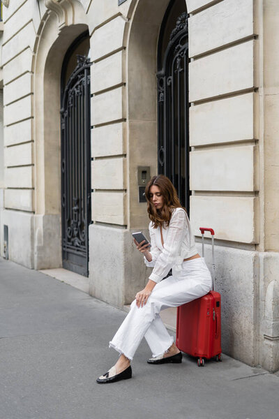 Brunette tourist using smartphone while sitting on suitcase on urban street in Paris 