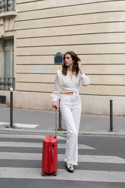 Trendy woman with suitcase walking on crosswalk on urban street in Paris