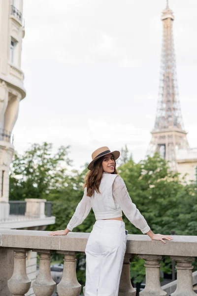 Happy young woman in blouse and sun hat standing with Eiffel tower at background in Paris