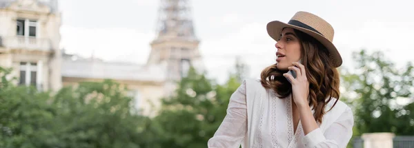 Woman in sun hat talking on smartphone with blurred Eiffel tower at background in Paris, banner