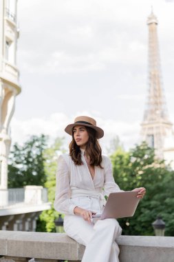 Şehir caddesinde dizüstü bilgisayar kullanan, güneş şapkalı, modaya uygun, Paris 'te arka planda Eiffel kulesi olan bir serbest yazar. 
