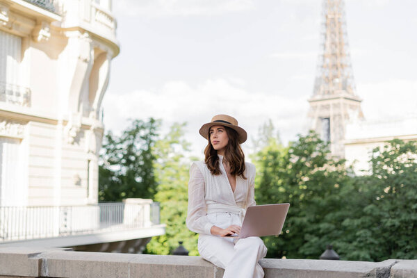 Trendy stylish freelancer in sun hat using laptop on street with Eiffel tower at background in Paris 