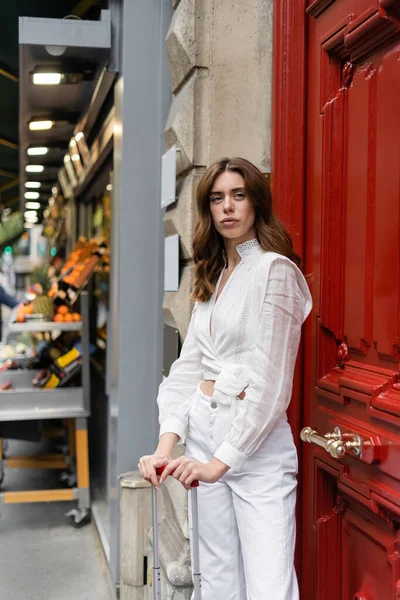 Woman in blouse standing near suitcase and door of building on urban street in Paris