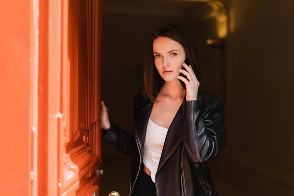 Young woman in black leather jacket talking on smartphone near orange door — Stock Photo