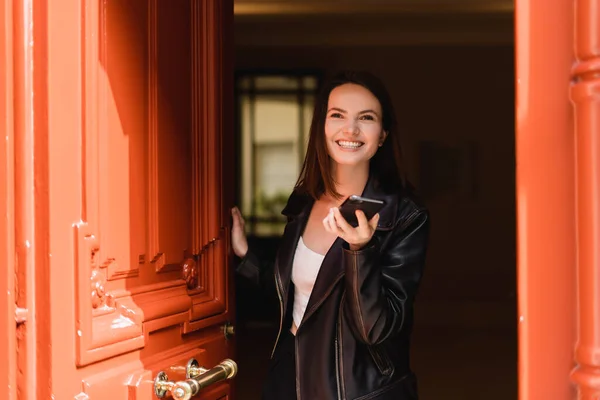 Happy young woman in black leather jacket holding smartphone near orange door — Stock Photo