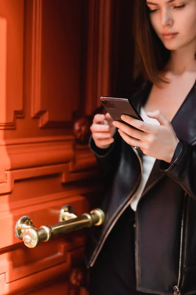 Giovane donna in giacca di pelle nera messaggistica su smartphone vicino alla porta arancione — Foto stock