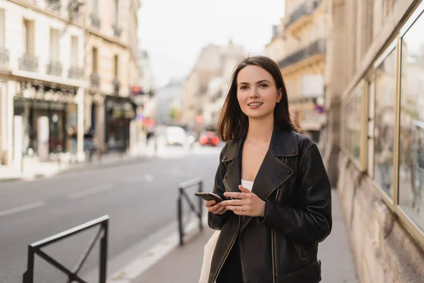 Allegra giovane donna in giacca di pelle nera con smartphone su strada a Parigi — Foto stock