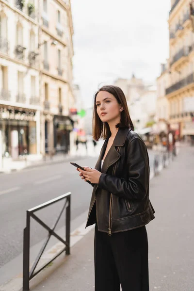 Jovem mulher em jaqueta de couro preto segurando smartphone na rua em Paris — Fotografia de Stock