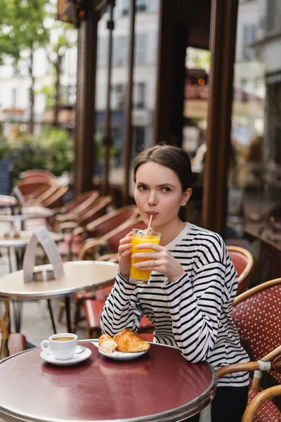 Jeune femme buvant du jus d'orange près du croissant et du café sur la table en terrasse d'été dans un café français — Photo de stock