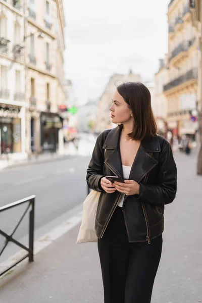 Mujer joven con chaqueta negra sosteniendo teléfono inteligente en la calle en París - foto de stock