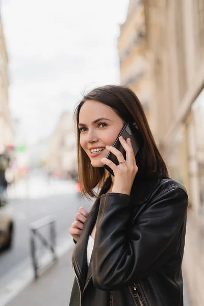 Позитивная молодая женщина в стильной куртке разговаривает по смартфону на улице в Париже — стоковое фото