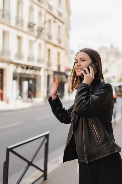 Happy woman in stylish jacket talking on smartphone and waving hand on street in paris — Stock Photo