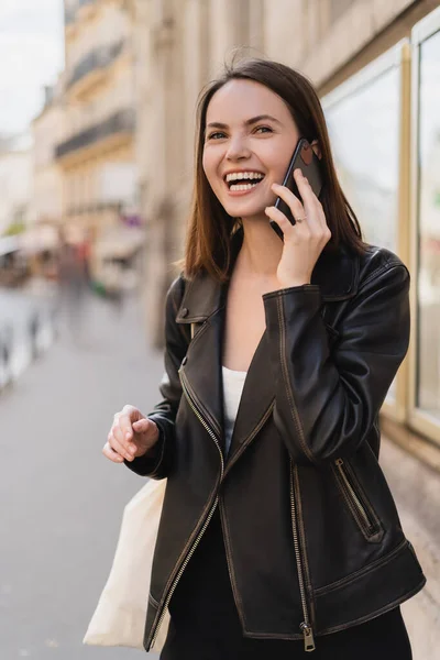 Happy young woman in stylish jacket talking on smartphone and laughing on street in paris — Stock Photo