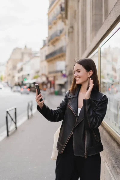 Heureuse jeune femme veste élégante agitant la main tout en ayant chat vidéo dans la rue à Paris — Photo de stock