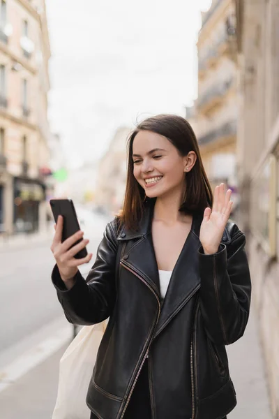 Cheerful young woman in stylish jacket waving hand during video chat on street in paris — Stock Photo