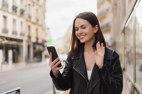 Mujer joven y complacida con elegante chaqueta saludando de la mano mientras tiene video chat en la calle en París - foto de stock