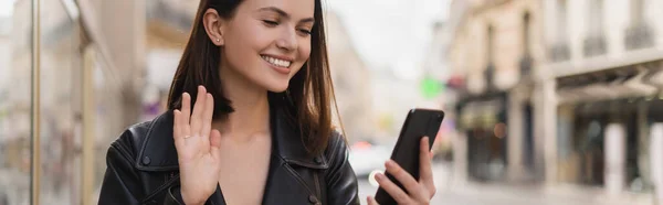 Jeune femme heureuse veste élégante agitant la main tout en ayant chat vidéo dans la rue à Paris, bannière — Photo de stock