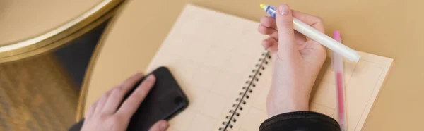 Vista recortada de la mujer que sostiene el rotulador y el teléfono inteligente cerca del cuaderno en blanco en la mesa redonda, pancarta - foto de stock