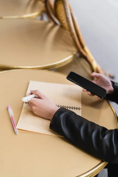 Cropped view of woman holding marker pen near blank notebook on round table and holding smartphone — Stock Photo