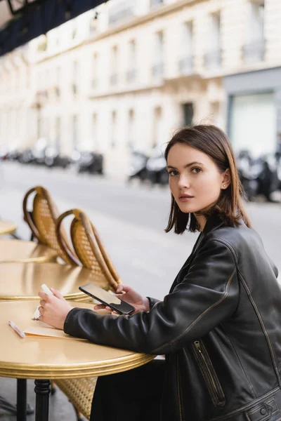 Young woman in leather jacket holding smartphone near notebook on round table in outdoor cafe terrace — Stock Photo